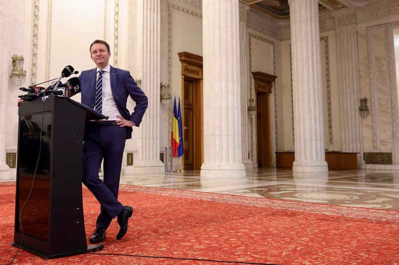 Premierul desemnat, europarlamentarul PNL, lângă un pupitru cu microfoane, într-un hol cu coloane de marmură