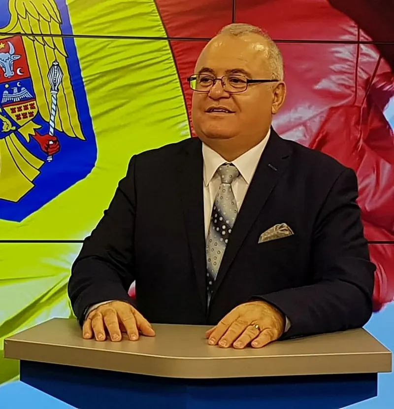 Ion Duvac, cu ochelari, în costum, la masa unui platou de televiziune, cu tricolorul în fundal