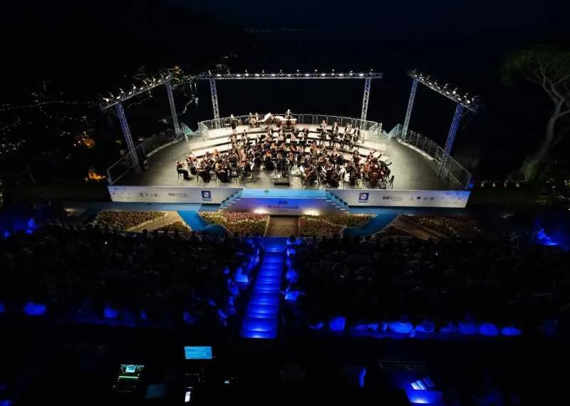 Orchestră pe o scenă în aer liber luminată noaptea, în fața publicului