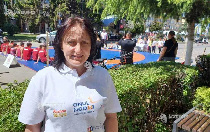 Femeie cu părul șaten, în tricou alb cu sigla „Anul Nadia”, lângă un spațiu cu saltele de gimnastică