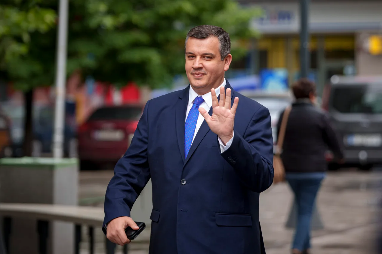 Bărbat în costum albastru făcând cu mâna pe o stradă, cu mașini parcate în spate