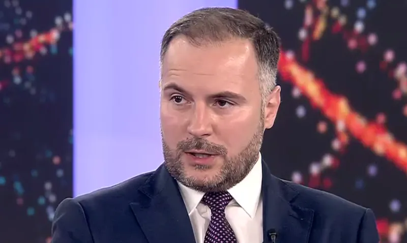 Bărbat cu barbă, în costum și cravată, vorbind într-un studio de televiziune