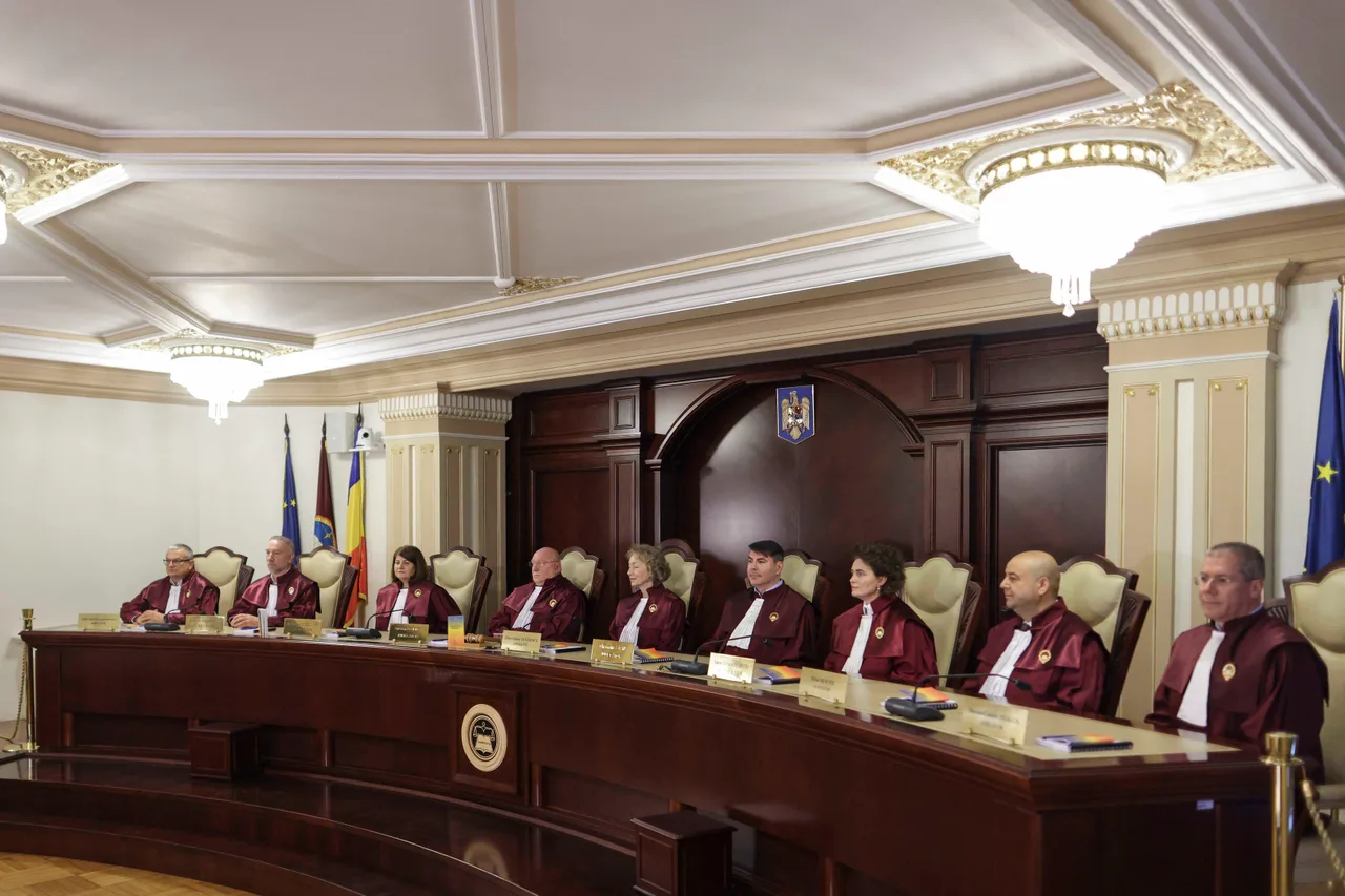 Judecători în robe violet, așezați la o masă lungă, într-o sală cu stema României și drapele