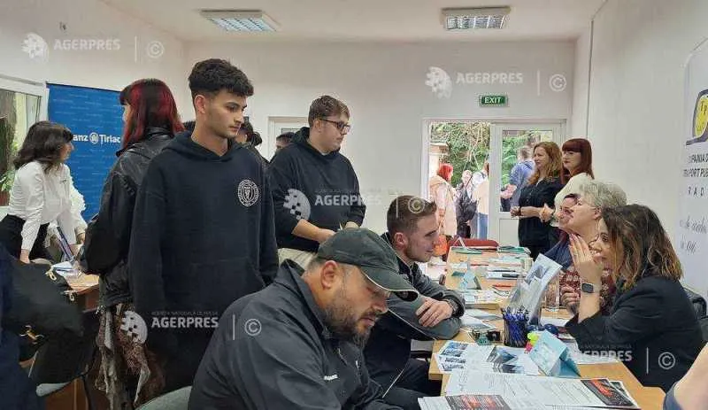 Tineri în jurul unor mese cu pliante, într-o sală aglomerată, vorbind cu oameni așezați