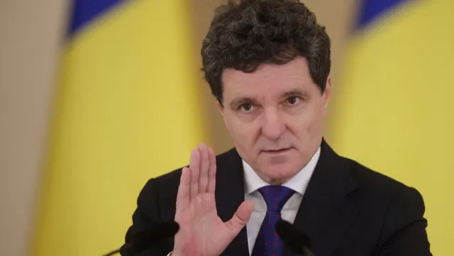 Bărbat cu părul creț, în costum și cravată mov, cu palma ridicată, în fața unor drapele galben-albastre