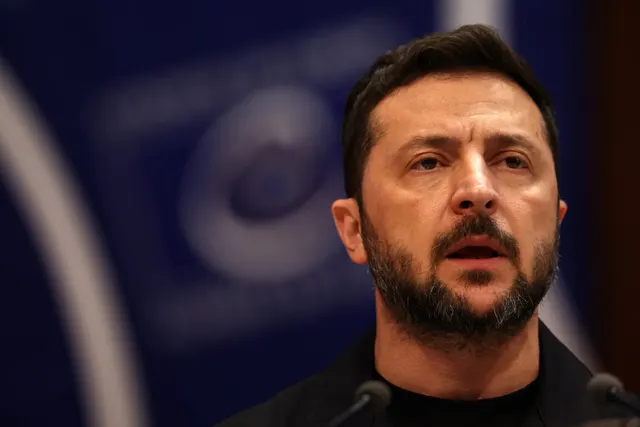 Volodimir Zelenski, în bluză neagră, vorbind în fața unui microfon, pe fundal albastru-închis