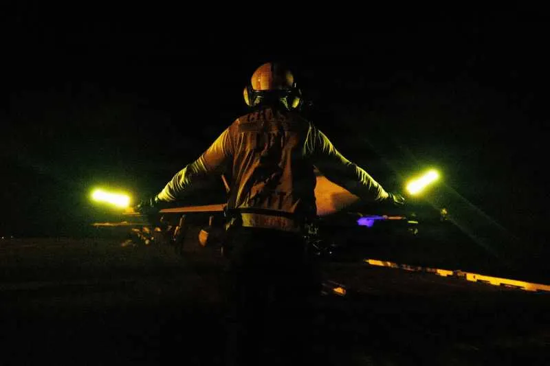 Membru al echipajului văzut din spate, noaptea, cu două baghete luminoase verzi ridicate pe puntea unei nave