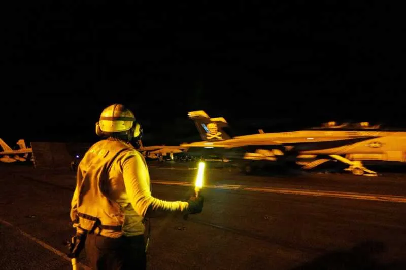 Membru al echipajului de punte cu baghete luminoase, ghidând noaptea un avion de luptă pe un portavion
