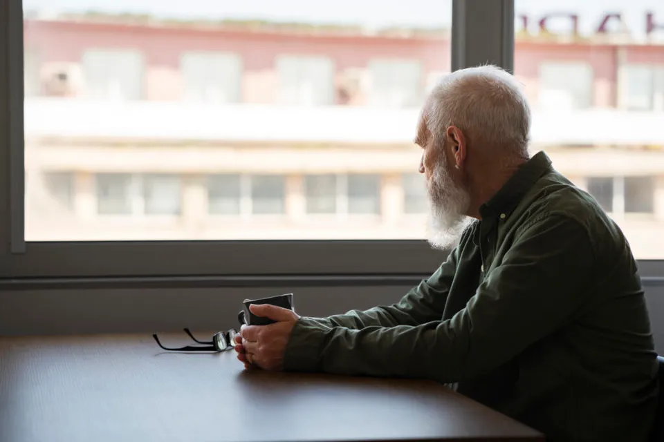 Bărbat în vârstă, cu barbă albă, stând singur la o masă lângă fereastră, cu o cană în față