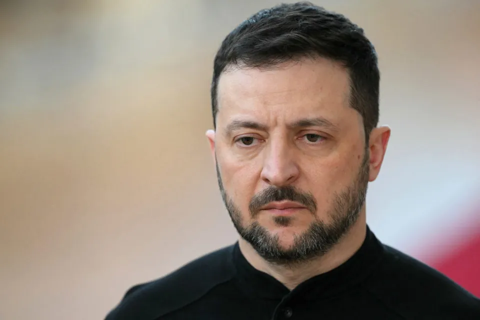 Volodimir Zelenski, cu barbă, în bluză neagră, privind spre aparat, pe fundal deschis