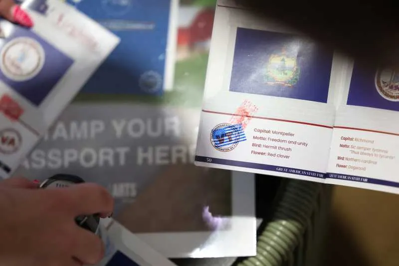 Mână lângă o broșură în formă de pașaport, cu un steag și inscripția „Stamp your passport here”