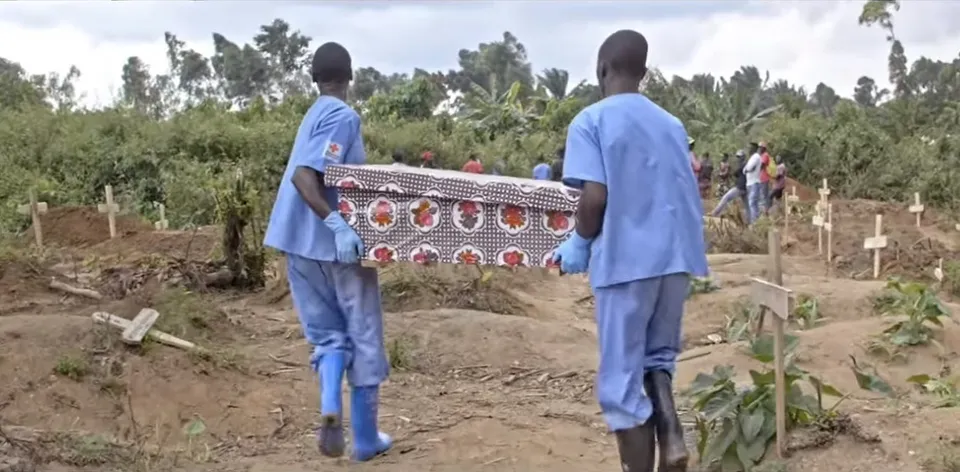 Doi oameni în echipament albastru purtând un sicriu printre cruci, la o înmormântare de Ebola din Congo