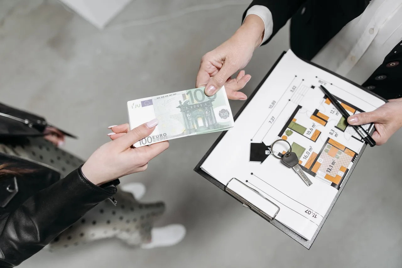 Mâini care schimbă o bancnotă de euro și chei, deasupra unui clipboard cu planul unei locuințe