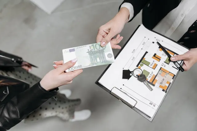 Mâini care schimbă o bancnotă de euro și chei, deasupra unui clipboard cu planul unei locuințe