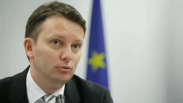 Bărbat în costum, cu părul pieptănat pe o parte, vorbind în fața unui drapel al UE