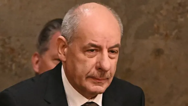 Bărbat în vârstă, chel, cu mustață, în costum închis, privind în jos
