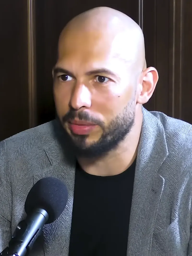 Andrew Tate, chel, cu barbă, în sacou gri, vorbind la un microfon