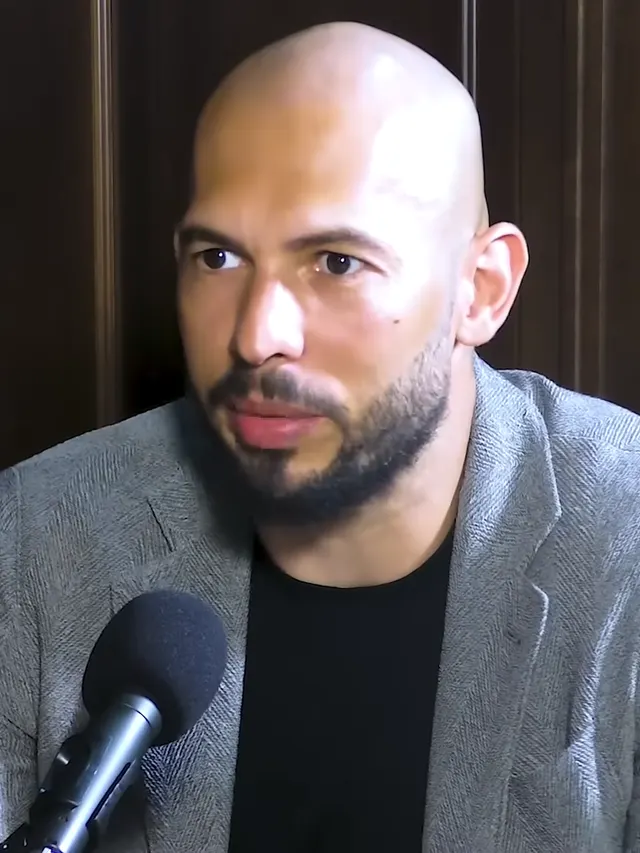 Andrew Tate, chel, cu barbă, în sacou gri, vorbind la un microfon