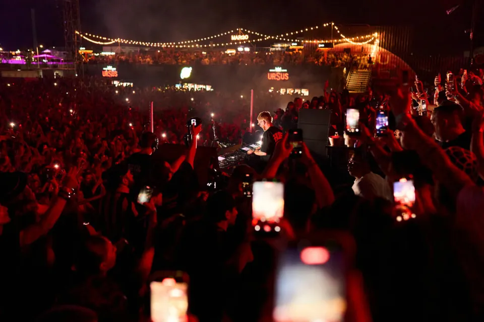 Mulțime la un festival noaptea, cu telefoane ridicate și lumini roșii deasupra scenei