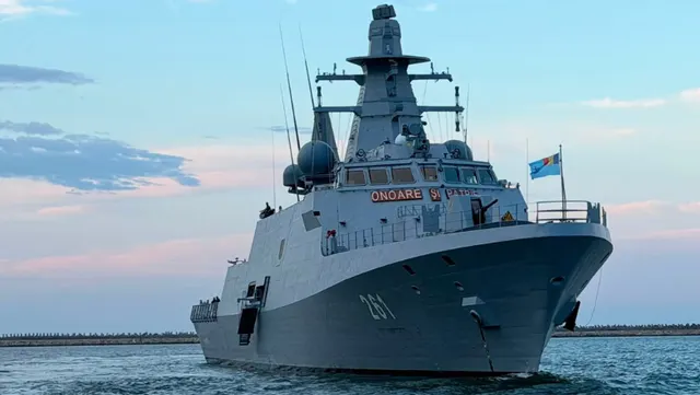 Navă militară gri, de tip corvetă, navigând pe mare sub un cer cu nori rozalii