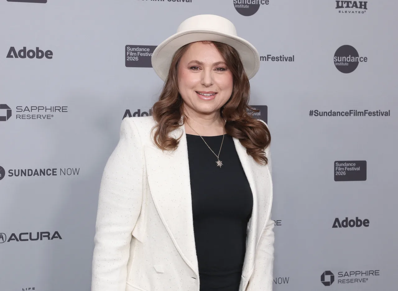 Femeie cu pălărie albă și sacou alb, la Sundance Film Festival, în fața panoului cu sponsori