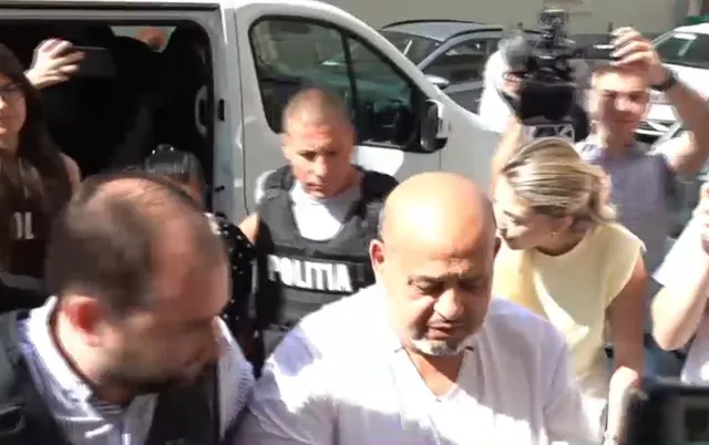 Polițiști în veste escortând doi bărbați printre jurnaliști și camere de filmat, lângă o dubă