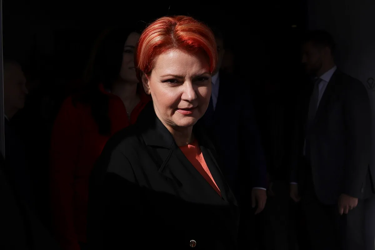 Femeie cu părul roșcat, în sacou închis, într-un loc întunecat, cu oameni în spate
