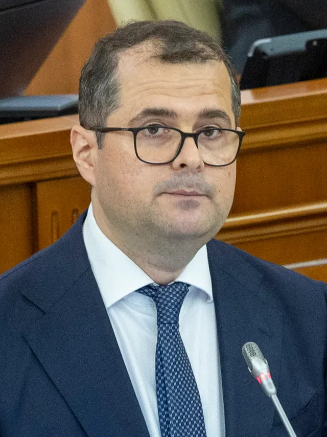 Vasile Tofan, cu ochelari și costum închis, la un microfon într-o sală de ședințe