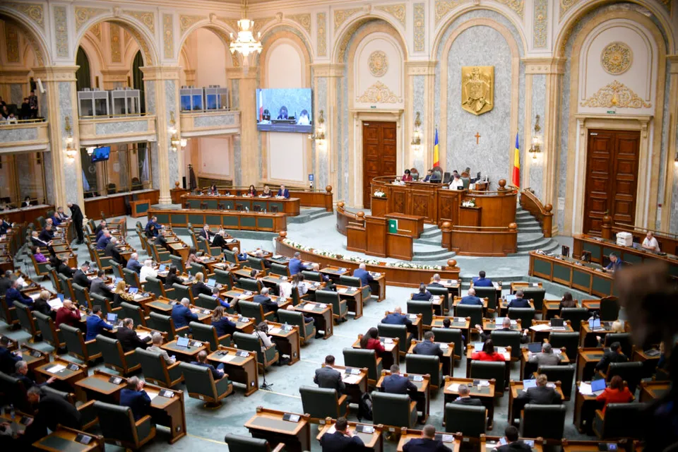 Sală de plen ornamentată, cu parlamentari în bănci și o stemă aurie pe peretele din fundal