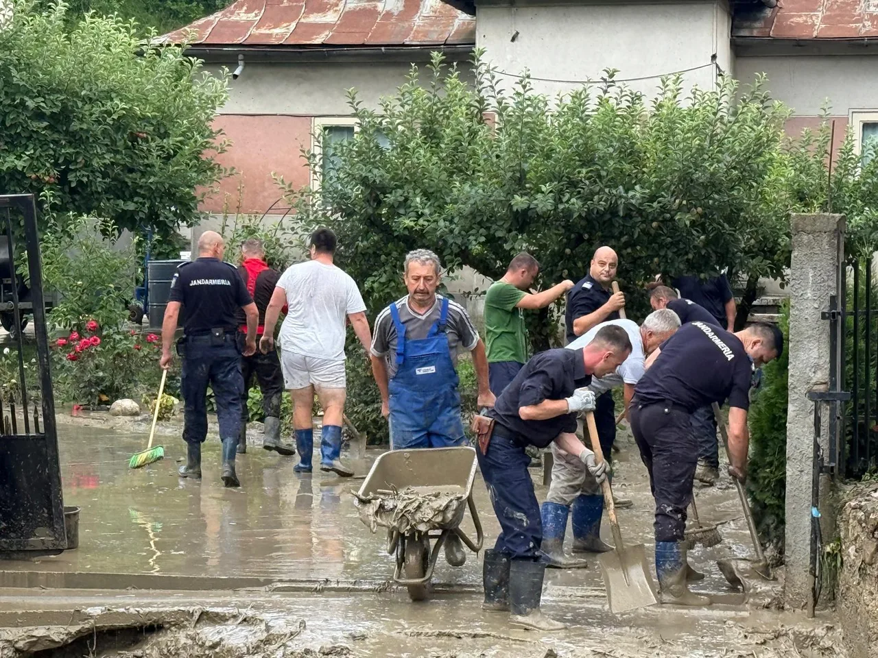 Pompieri și localnici cu lopeți și o roabă, curățând noroiul dintr-o curte după inundații