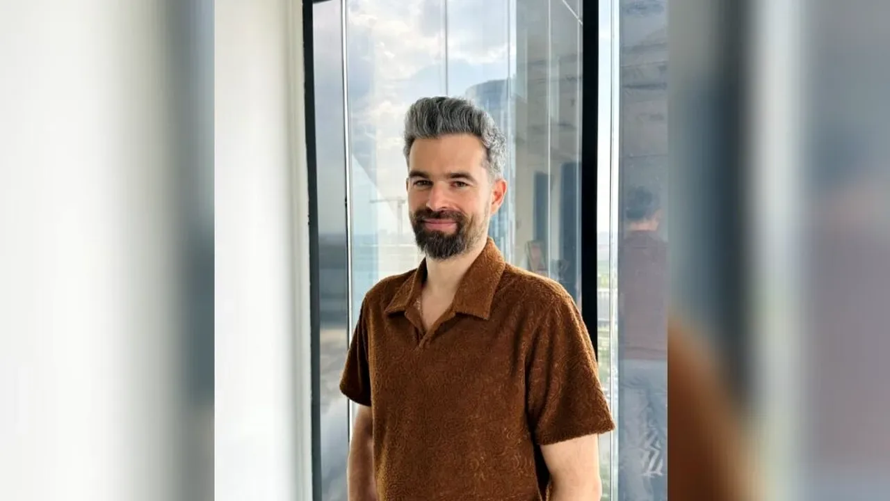 Bărbat cu barbă și părul grizonat, în cămașă maro, zâmbind lângă o fereastră