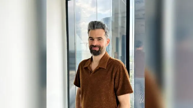 Bărbat cu barbă și părul grizonat, în cămașă maro, zâmbind lângă o fereastră