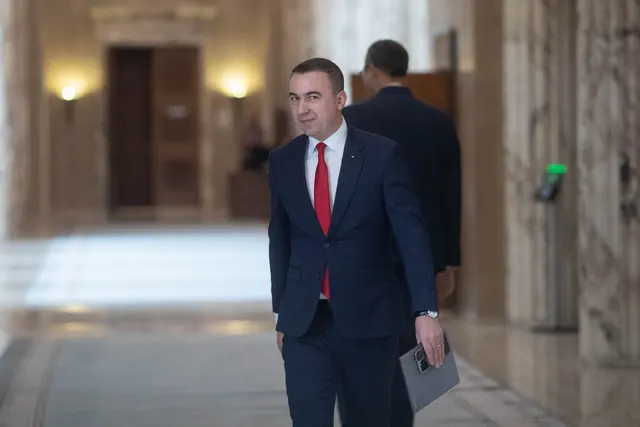 Bărbat în costum și cravată roșie mergând pe un hol de marmură, cu un dosar în mână
