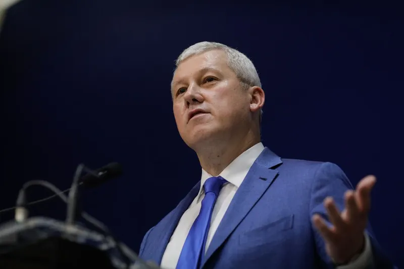 Bărbat cu părul alb, în costum albastru, gesticulând cu mâna întinsă în timp ce vorbește
