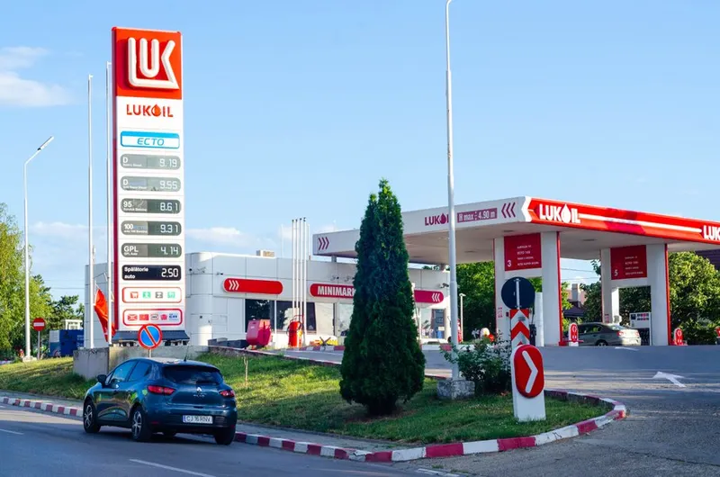Benzinărie Lukoil cu panoul de prețuri și copertina roșie, cu o mașină albastră pe aleea de acces