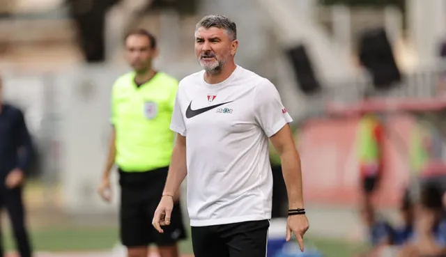 Bărbat în tricou alb, pe marginea terenului, lângă un arbitru în echipament galben-verzui