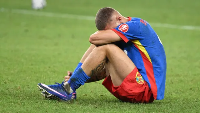 Fotbalist în echipament roș-albastru, așezat pe gazon cu capul plecat și brațele pe genunchi