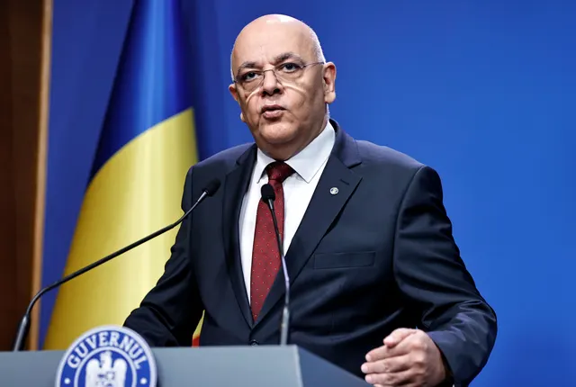 Bărbat chel, cu ochelari, în costum și cravată roșie, vorbind la pupitrul Guvernului României