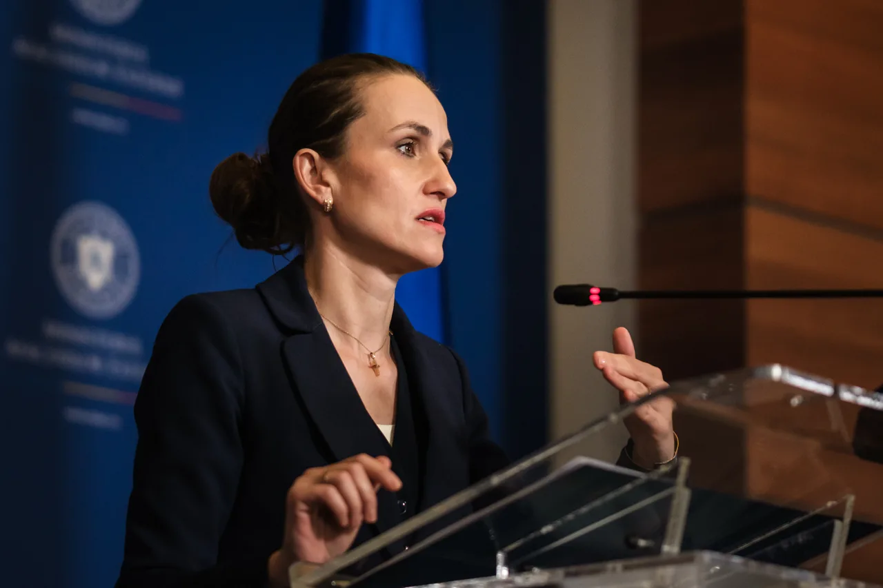 Femeie cu părul prins în coc, în sacou închis, vorbind la un pupitru de sticlă cu microfon