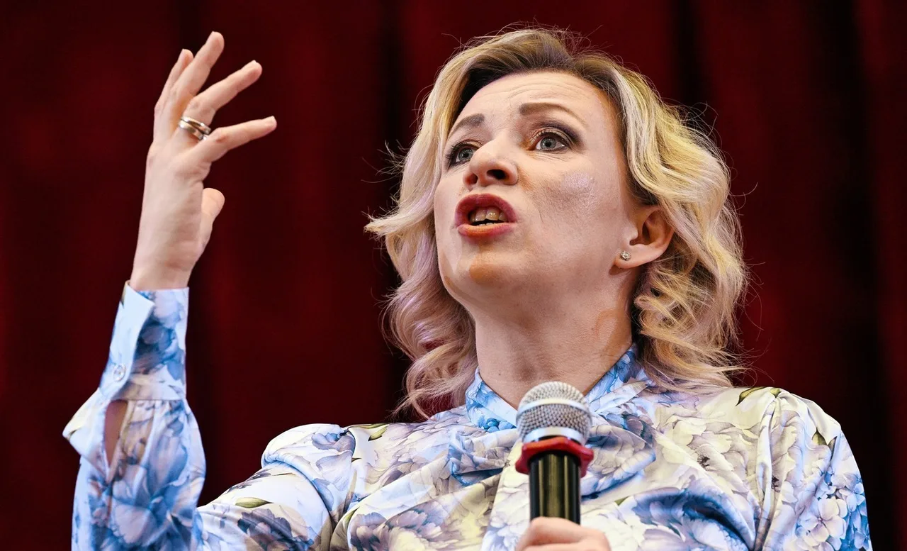 Femeie blondă, în bluză albastră cu model, vorbind la microfon cu mâna ridicată, în fața unei cortine roșii