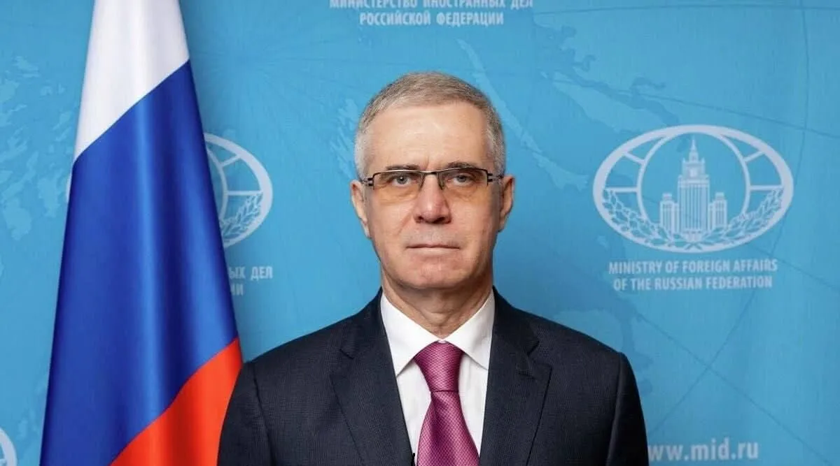 Bărbat cu ochelari, în costum, lângă drapelul Rusiei, în fața panoului Ministerului de Externe rus