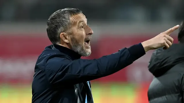 Bărbat cu barbă grizonată, în geacă închisă, arătând cu degetul și strigând, la un meci