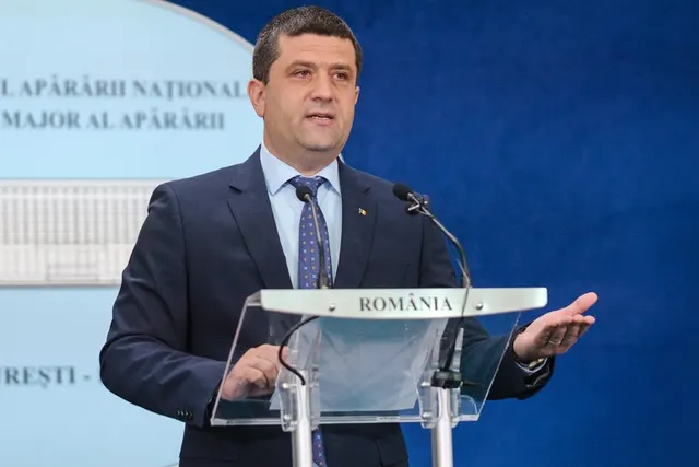Bărbat în costum la un pupitru cu inscripția „România”, în fața unui panou al Ministerului Apărării Naționale