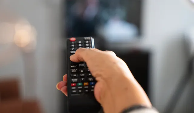 Mână care ține o telecomandă îndreptată spre un televizor, într-o cameră