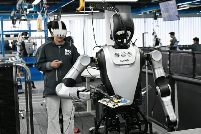 Robot umanoid manipulând o placă electronică, ghidat de un om cu cască de realitate virtuală
