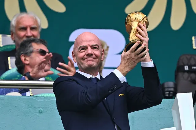Bărbat chel, în costum, ridicând trofeul Cupei Mondiale într-o tribună, lângă alți oficiali