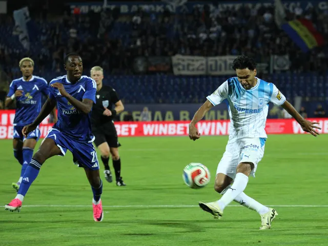 Fotbalist în echipament alb șutând, urmărit de adversari în albastru, în fața panourilor Winbet