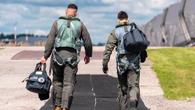 Doi piloți militari în combinezoane de zbor, cu genți de cască, mergând pe platforma unei baze aeriene