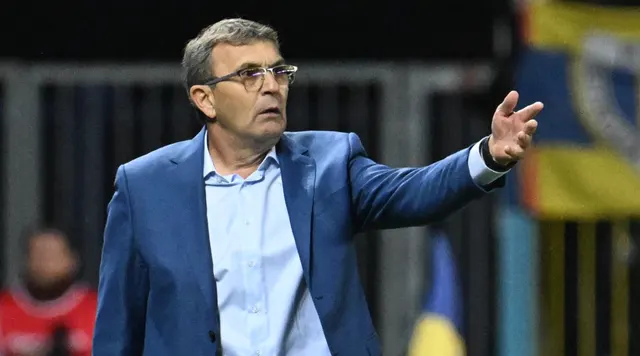 Bărbat cu ochelari, în sacou albastru, gesticulând cu mâna întinsă pe marginea terenului