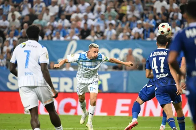 Fază aeriană între jucători în alb și un fundaș în albastru cu numărul 47, pe un stadion plin
